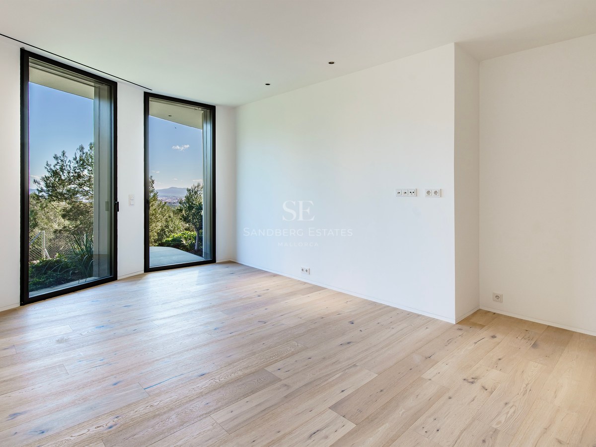 Une pièce vide avec du parquet en bois clair, des murs blancs et deux grandes baies vitrées donnant sur des arbres verts.