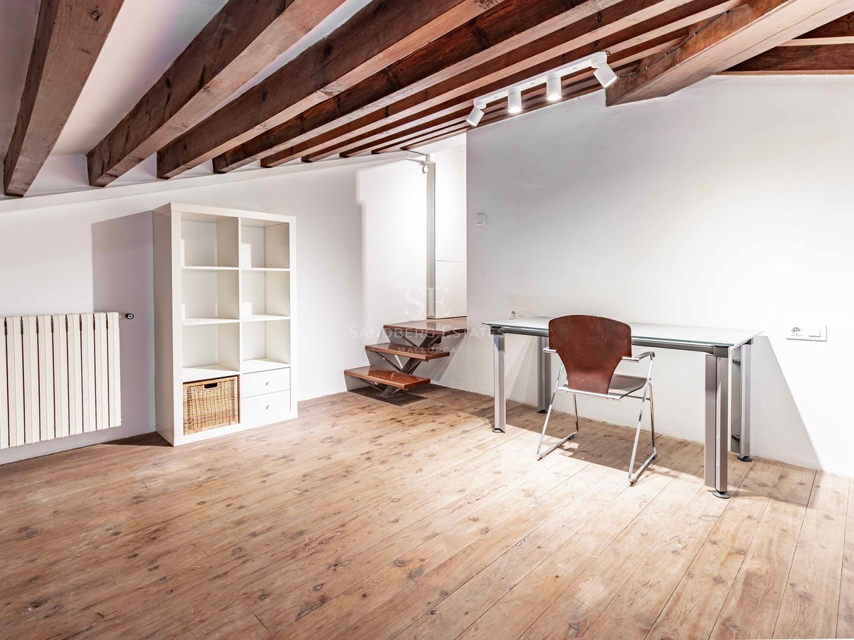 Attic office featuring exposed wooden beams, a glass desk, wooden floors, and a minimalist white shelf.