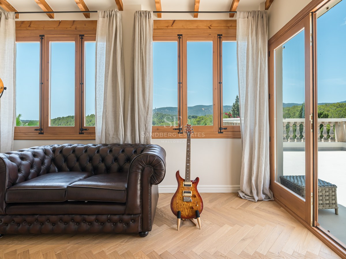 Living room with brown leather Chesterfield sofa, oak herringbone flooring, and large windows overlooking hills.