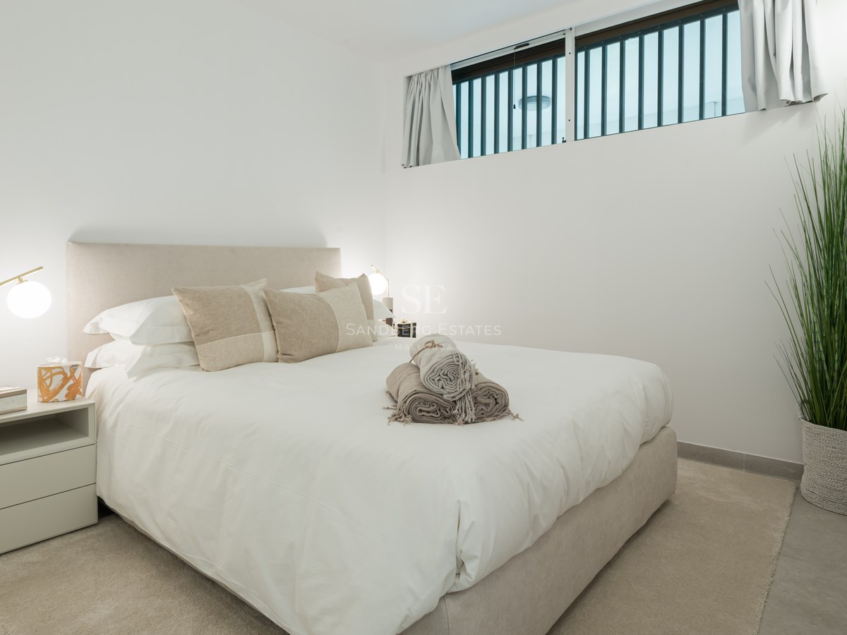 Bright bedroom with beige upholstered bed, white linens, brass lamps, and a tall indoor plant.