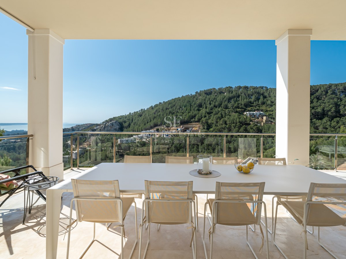 Terraza cubierta moderna con mesa de comedor blanca y barandillas de cristal con vistas a colinas verdes y al mar.