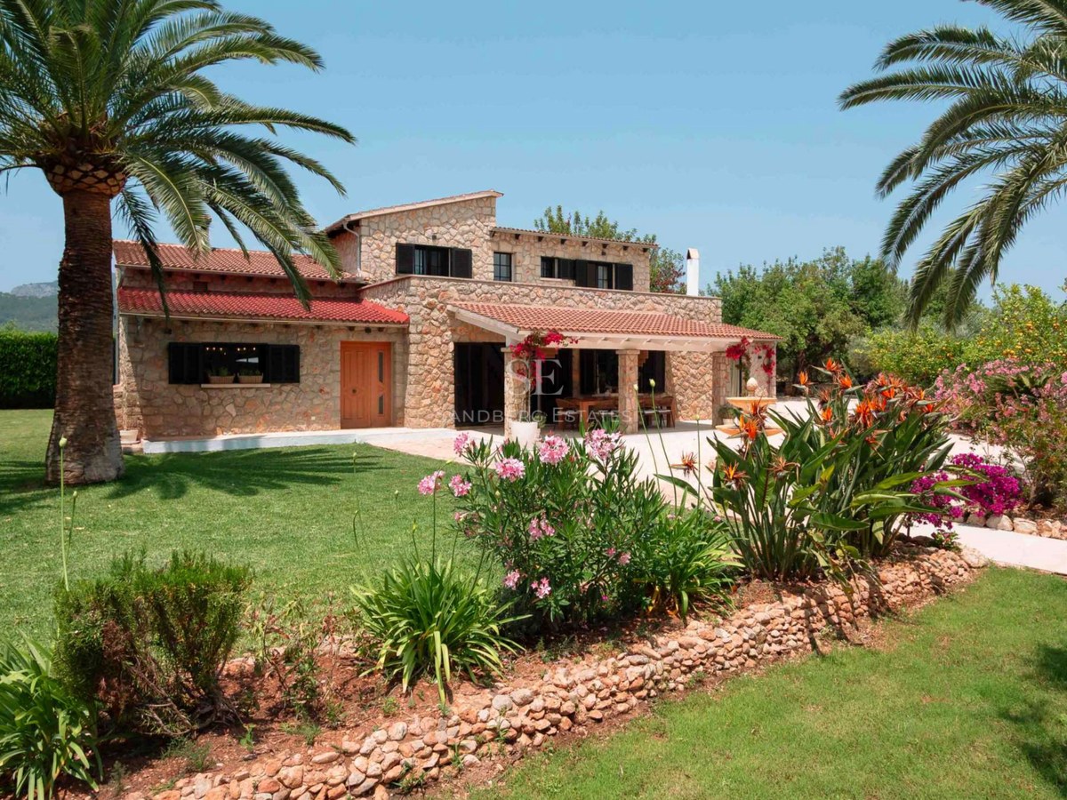 Two-story natural stone villa with terracotta roofs, lush lawn, palm trees, and exotic flowers under a clear blue sky.
