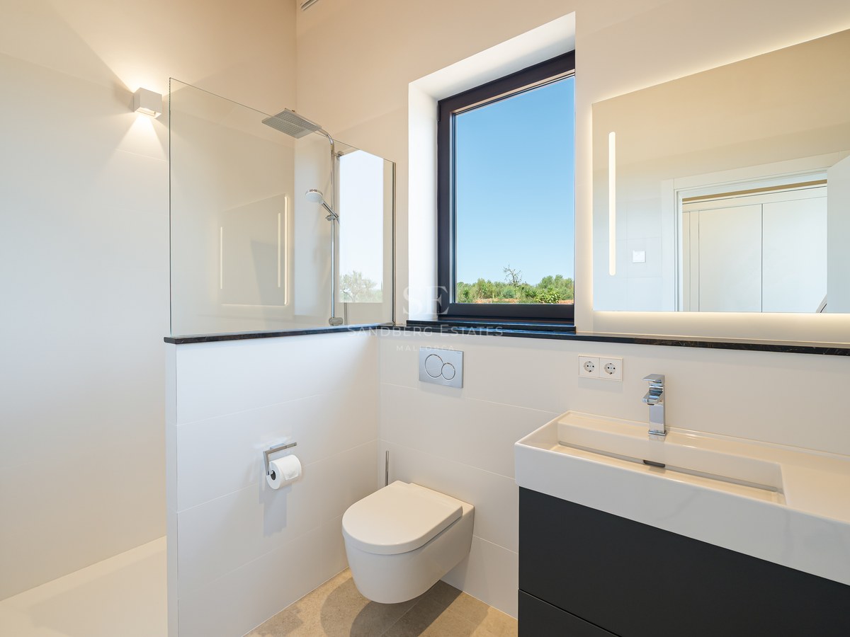 Minimalist white bathroom featuring a glass walk-in shower, wall-hung toilet, and dark vanity unit under a window.