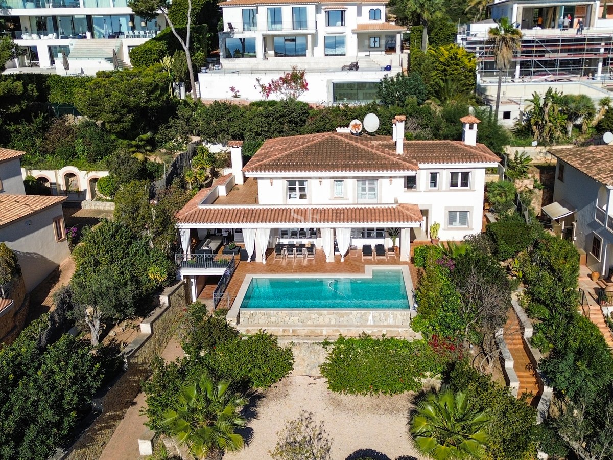 Vue aérienne d'une villa blanche à toit de tuiles avec piscine turquoise et jardins luxuriants.
