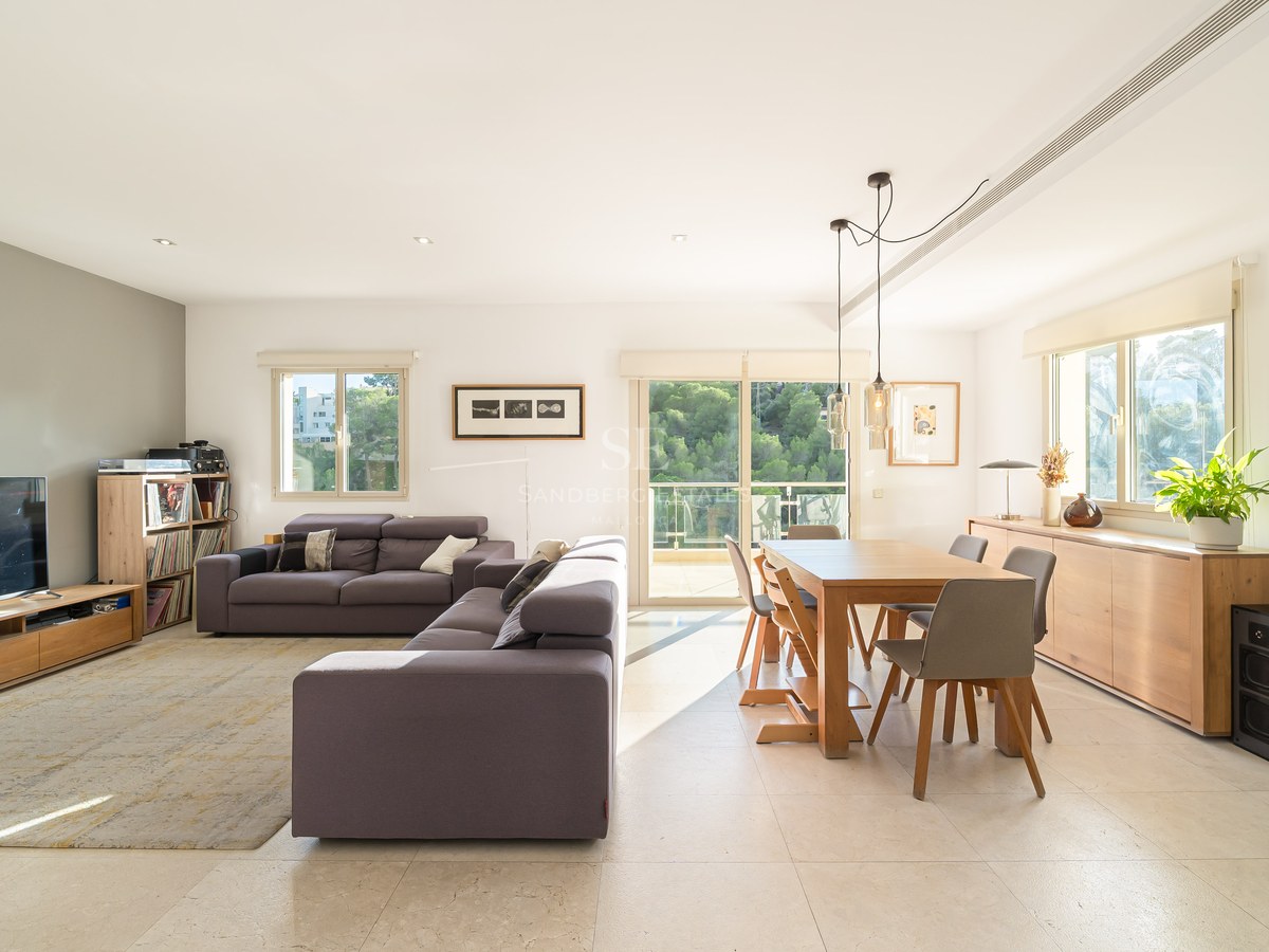 Bright living room featuring a grey sofa, wooden dining table, and large windows overlooking lush green trees.