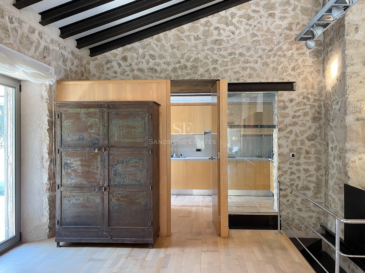Habitación interior con paredes de piedra natural, suelos de madera clara, vigas oscuras y vista a una cocina moderna.