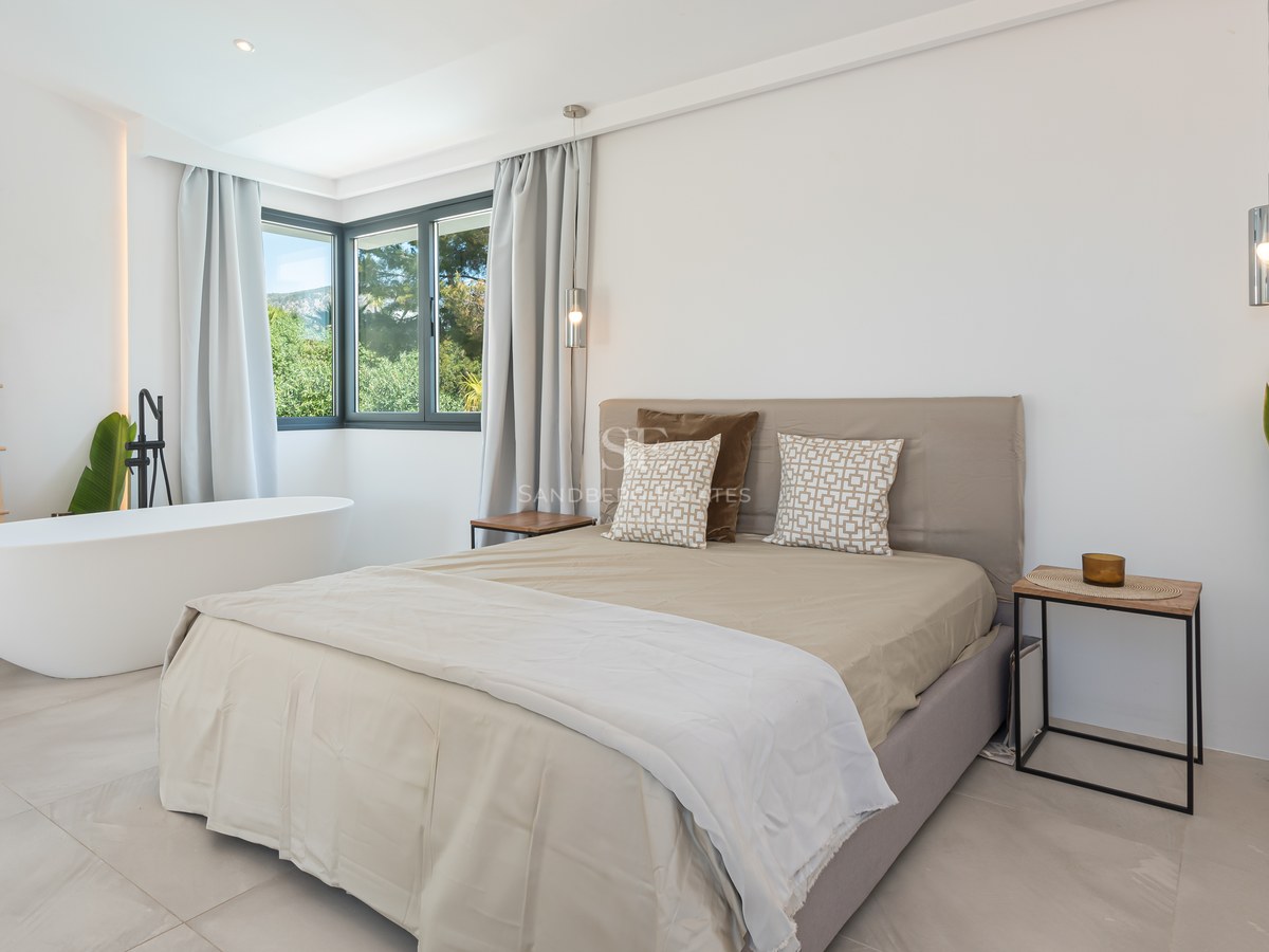 Minimalist bedroom featuring a white freestanding tub, neutral linens, and corner windows overlooking lush greenery.