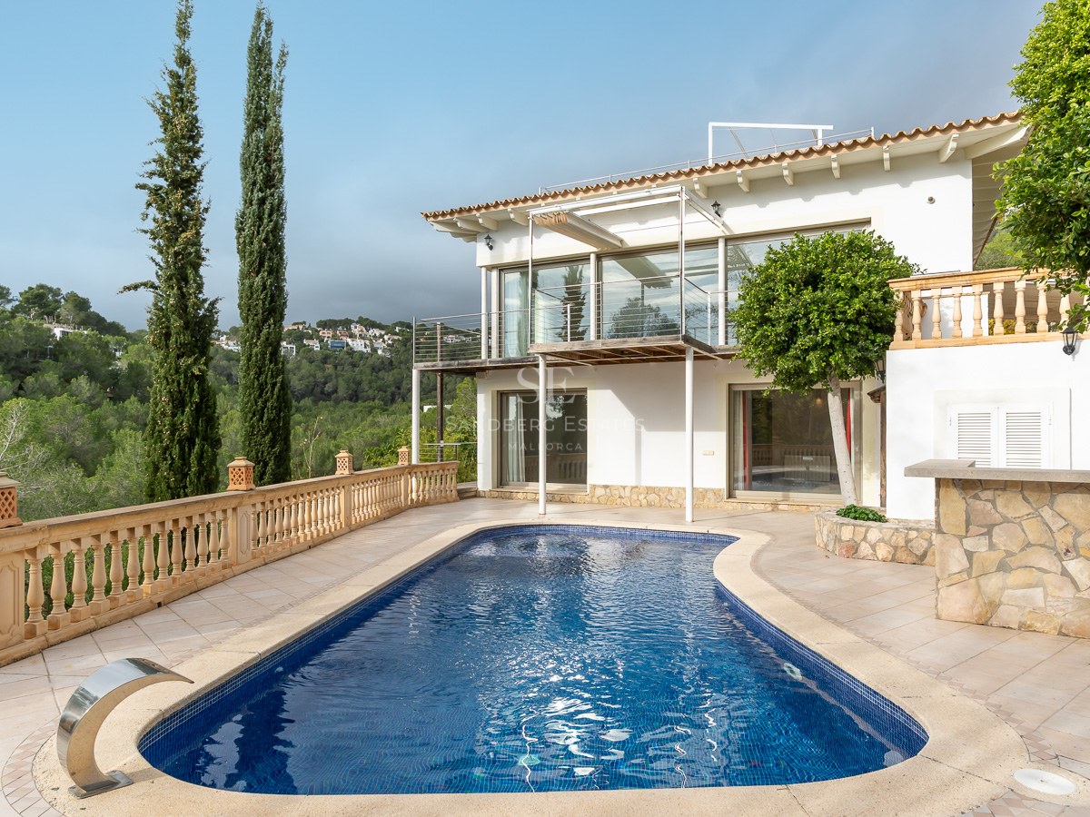 A kidney-shaped swimming pool on a stone terrace next to a two-story white villa overlooking a lush green forest.