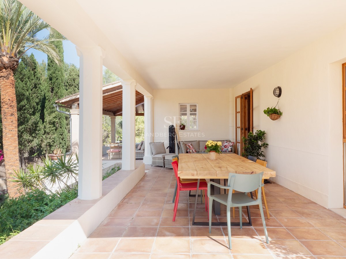 Covered terrace with terracotta floors, a large wooden dining table, and view of lush garden with palm trees.