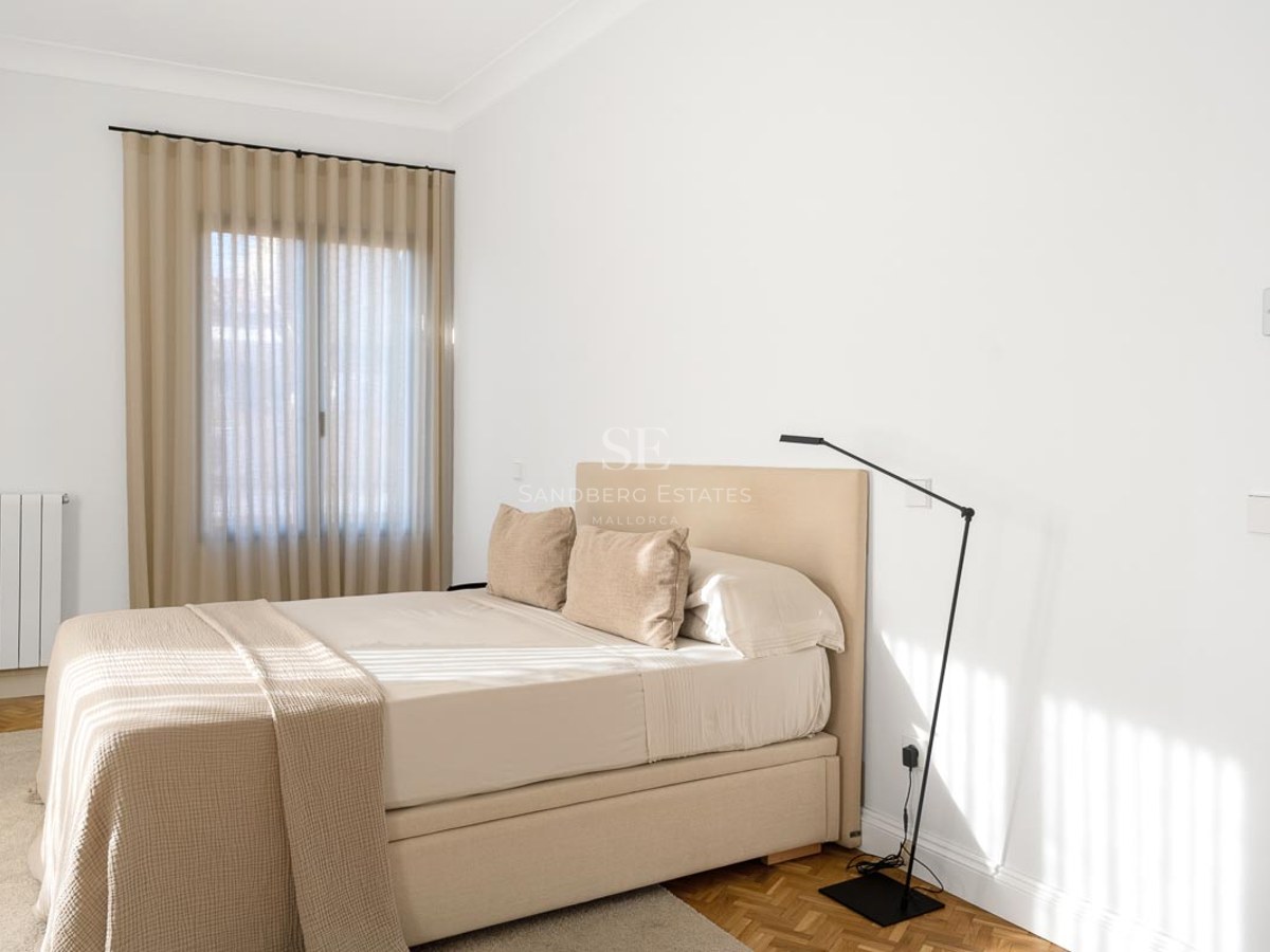 Bright minimalist bedroom with a beige upholstered bed, white walls, and warm wooden parquet flooring.