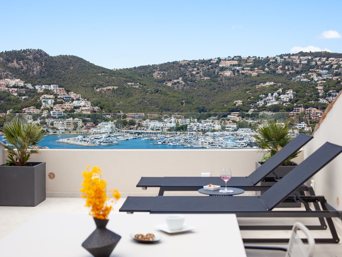 Sun loungers on a modern terrace overlooking a harbor with white boats and lush green hills under a clear blue sky.