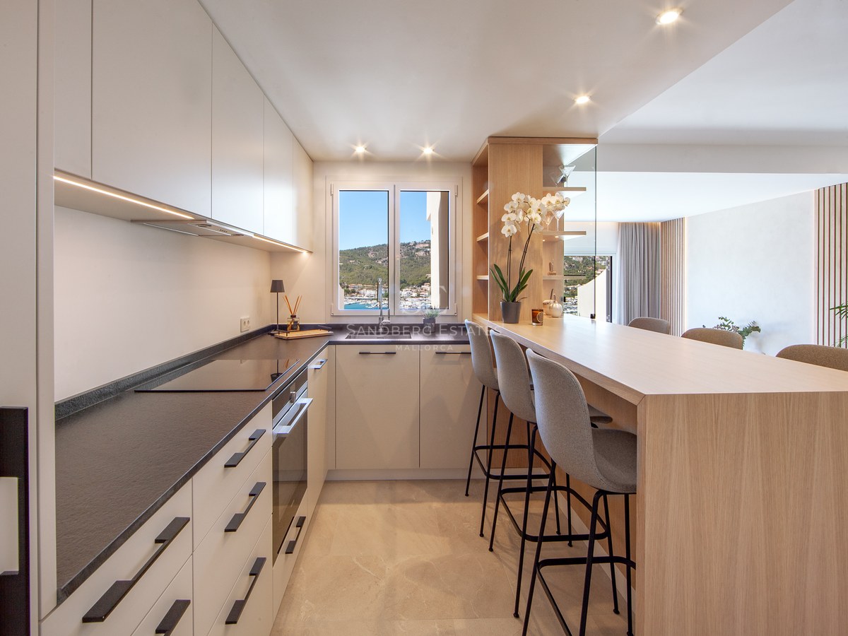 Modern kitchen with white cabinets, dark countertops, a wooden breakfast bar with three stools, and a window view of a harbor.