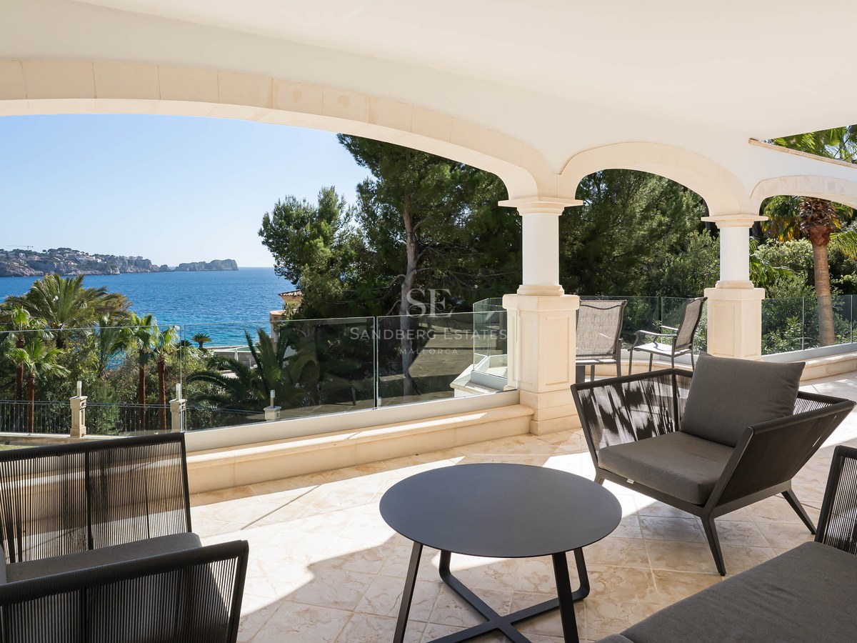 Covered stone terrace with arches, glass railings, and modern furniture overlooking the Mediterranean Sea and pine trees.