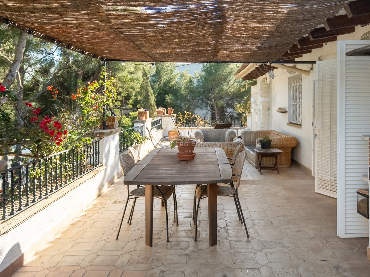 Terraza sombreada con mesa de comedor de madera, muebles de mimbre y barbacoa integrada rodeada de vegetación.