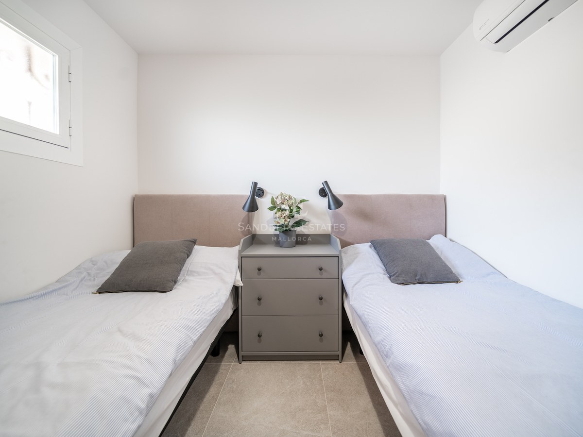 Chambre lumineuse avec deux lits simples, têtes de lit beiges, commode grise centrale et liseuses murales noires.
