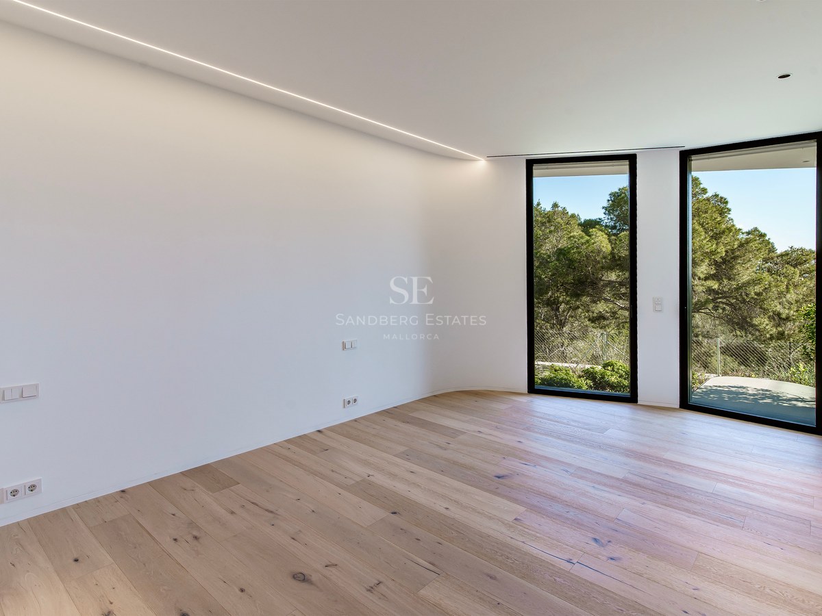 Pièce vide lumineuse avec parquet en bois clair, murs blancs et grandes fenêtres donnant sur la forêt.