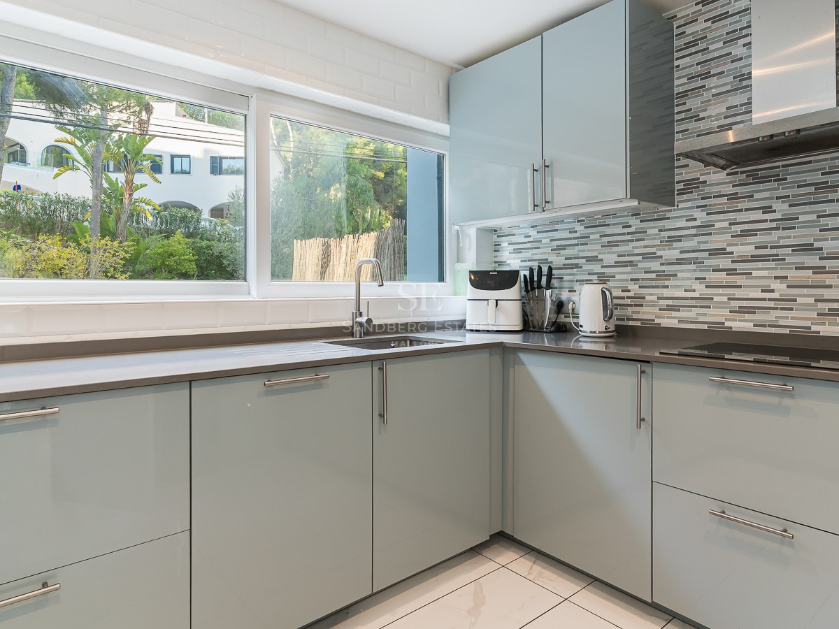 Modern light blue kitchen cabinets with grey countertops, a large window overlooking trees, and a mosaic tile wall.
