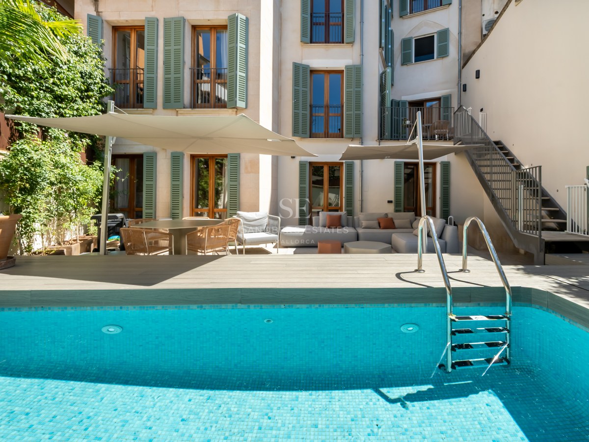 A turquoise swimming pool with a wooden deck, lounge furniture under shade sails, and a building with green shutters.