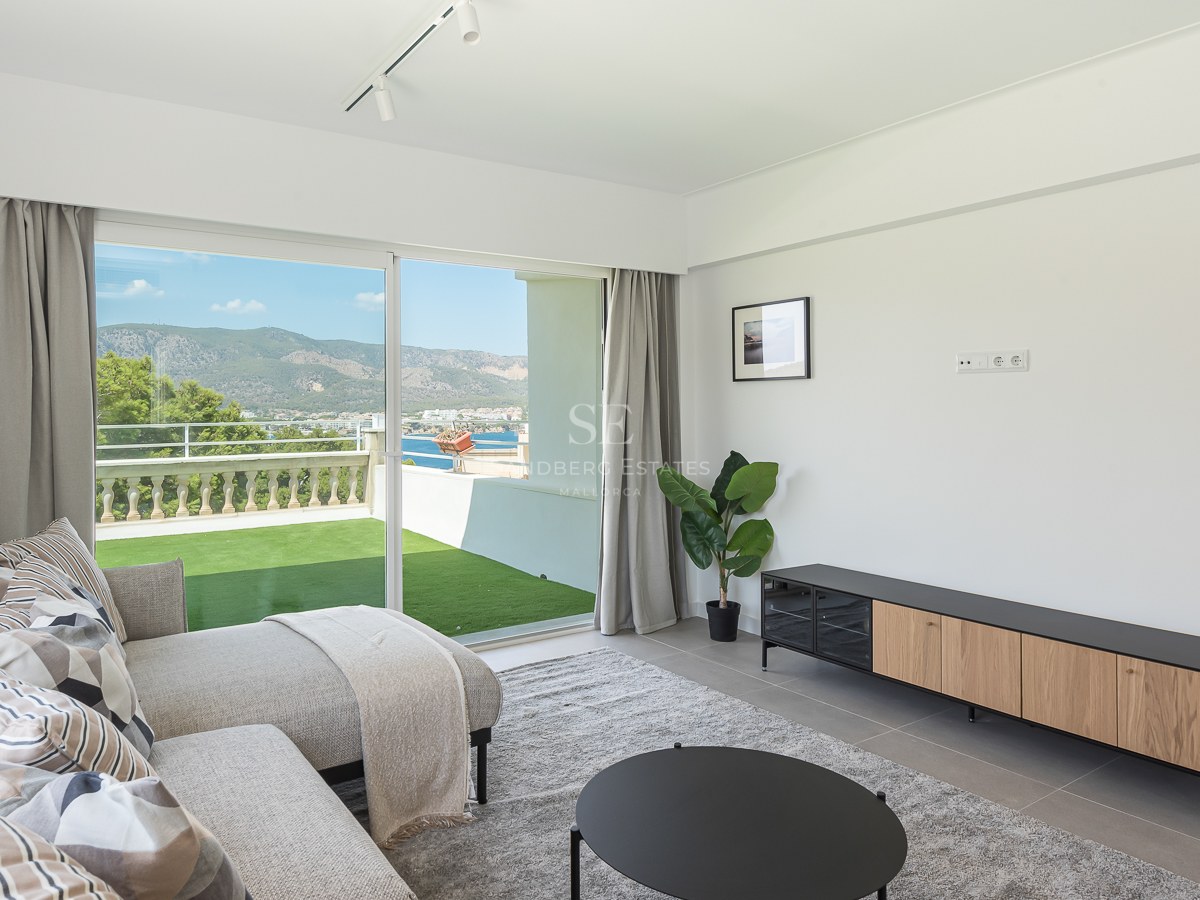 Salon moderne avec canapé gris et baies vitrées ouvrant sur une terrasse vue mer et montagne.