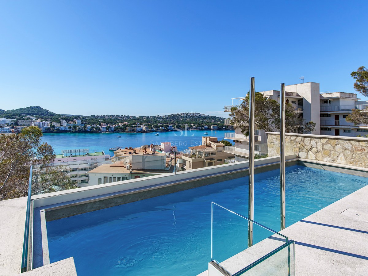 Piscine moderne avec garde-corps en verre surplombant une baie méditerranéenne et une ville côtière sous un ciel bleu.