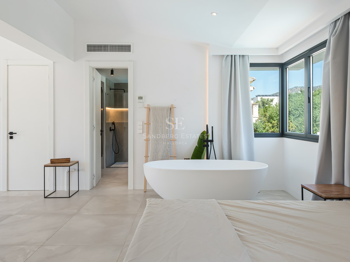 Large white bedroom featuring a freestanding oval bathtub, grey curtains, and an en-suite shower room in the background.