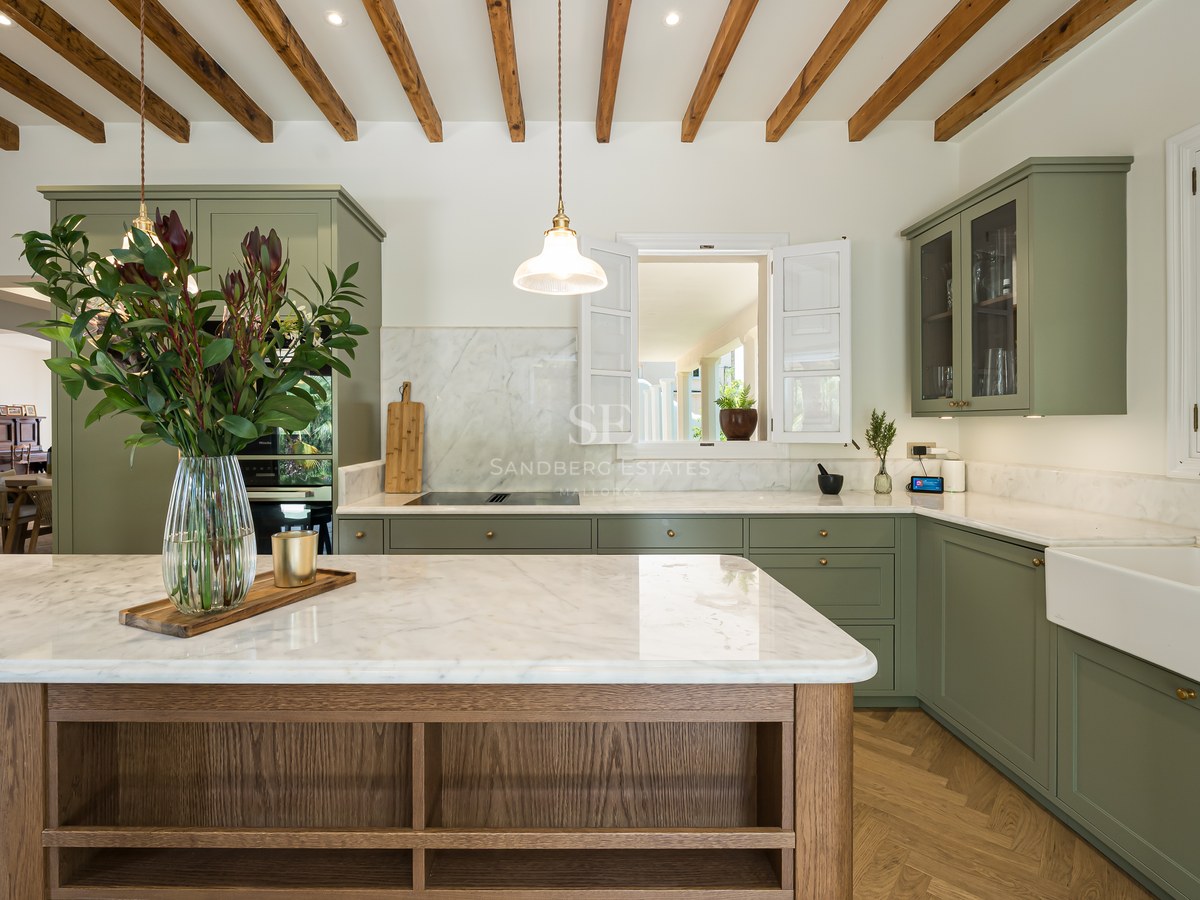 Modern sage green kitchen with marble countertops, wooden ceiling beams, and a large central island with flowers.