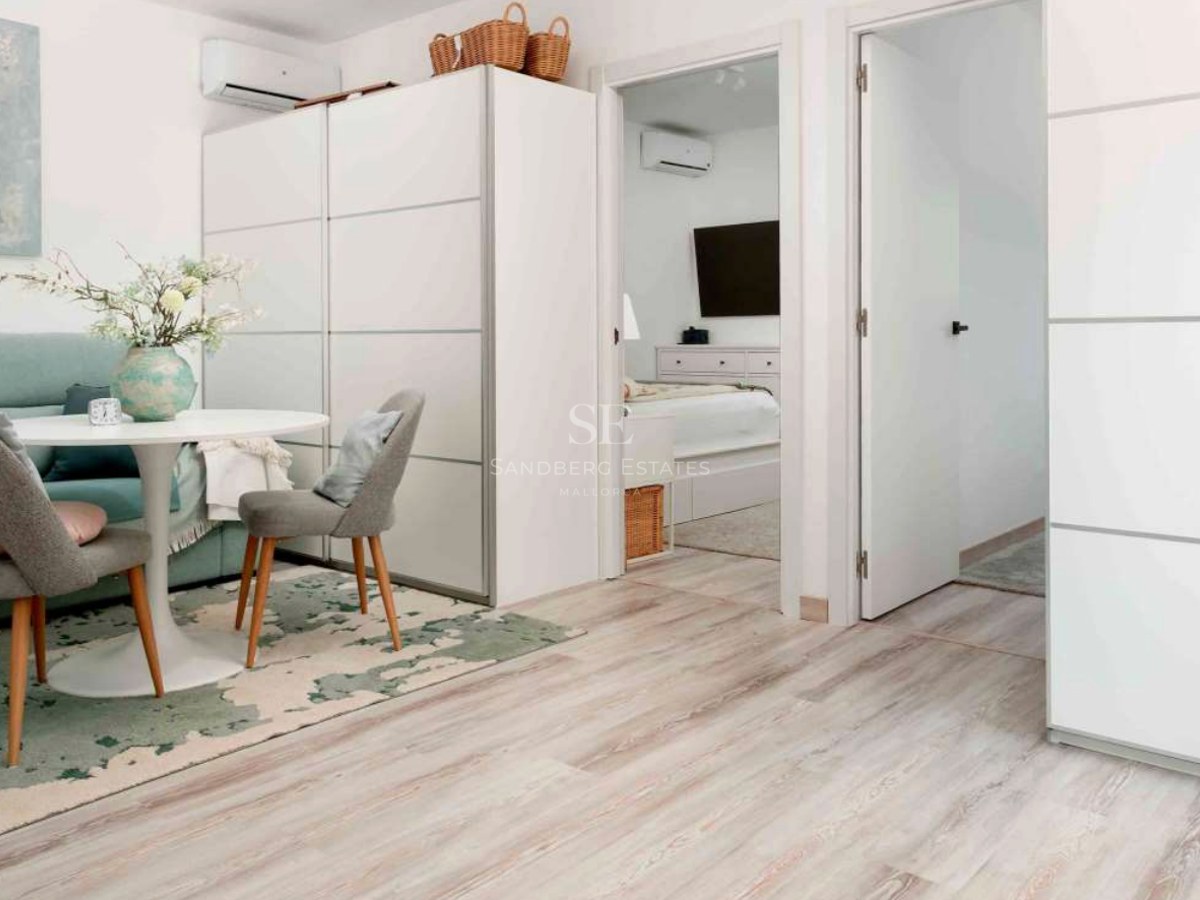 Bright living space featuring a round white table, grey chairs, light wood floors, and a view into an adjacent bedroom.