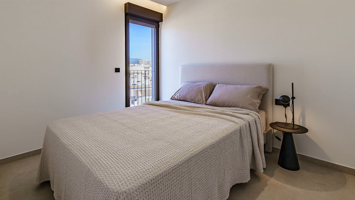 Chambre minimaliste avec lit double, linge de maison beige, lampe de chevet noire et porte vitrée vue ville.