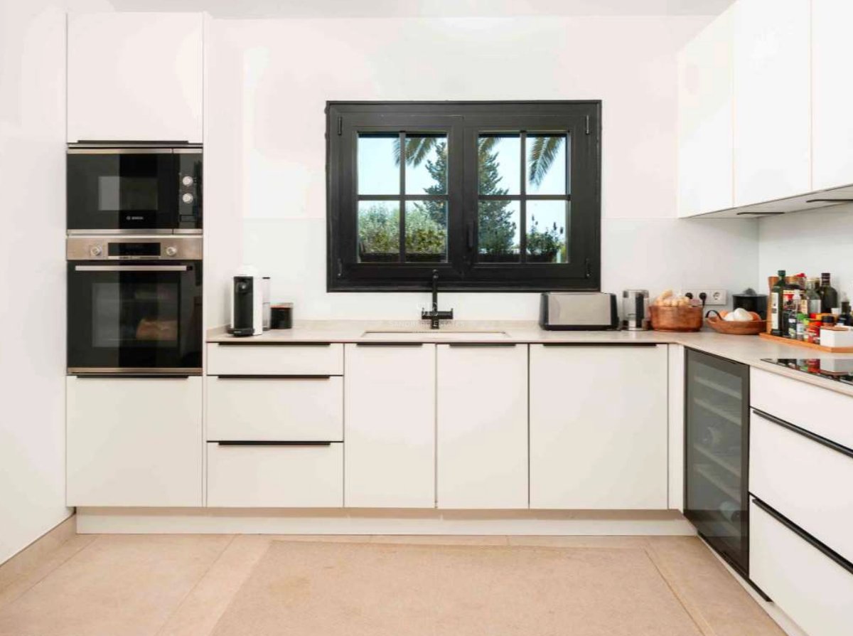 Cuisine blanche moderne avec fenêtre à cadre noir, four encastré, cave à vin et plans de travail en pierre claire.