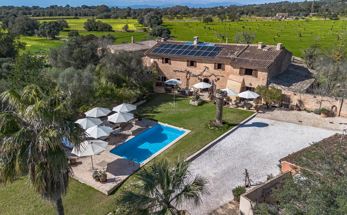 Vue aérienne d'une villa en pierre avec piscine bleue, panneaux solaires et parasols blancs entourée de champs.