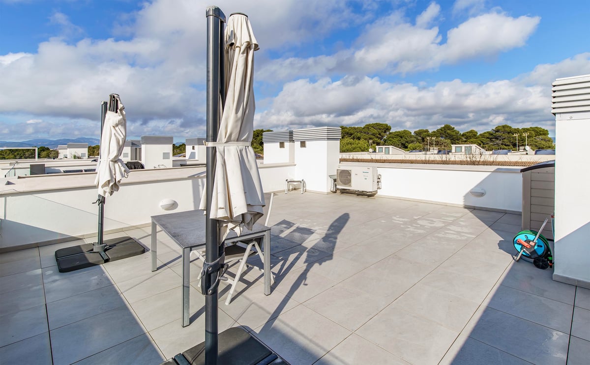 Grande terrasse sur le toit avec carrelage, table grise, chaises et parasols fermés sous un ciel nuageux.