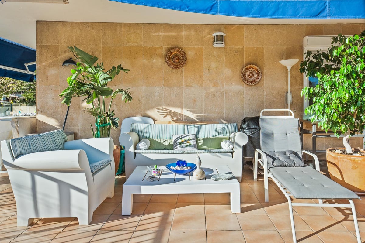 Terrasse ensoleillée avec mobilier de salon blanc, mur en pierre texturée et plantes vertes en pots.