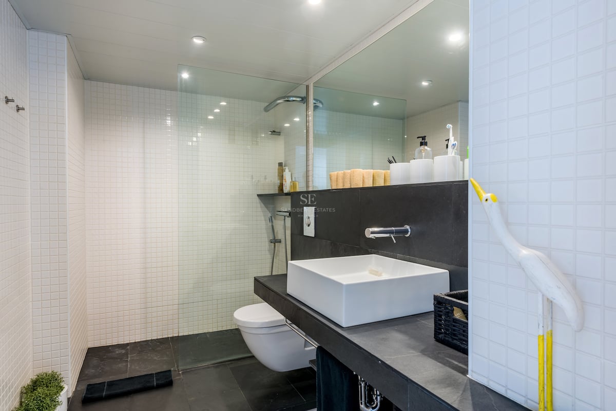 Salle de bain moderne avec carrelage blanc, sol noir, douche en verre et vasque carrée.