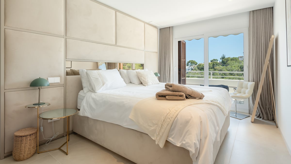 Chambre principale lumineuse avec tête de lit rembourrée, tables en verre et accès balcon vue jardin.