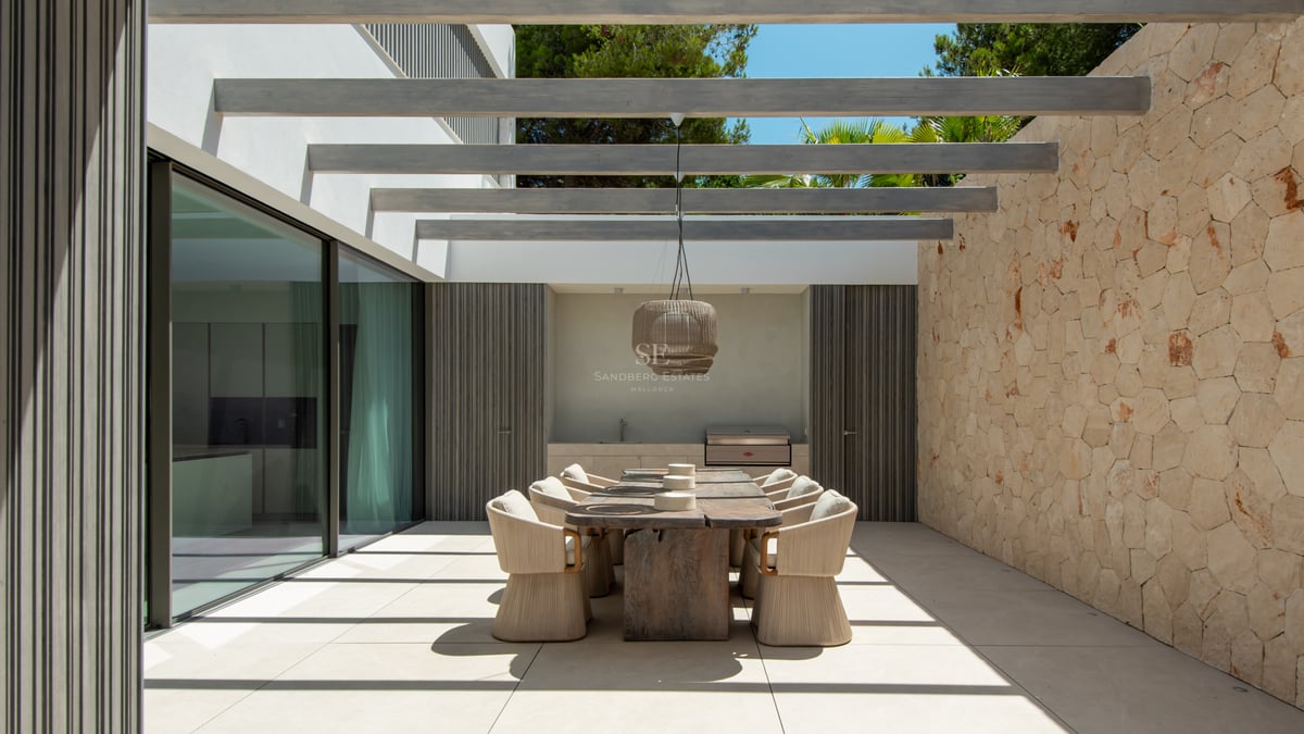 Terrasse moderne avec table en bois massif, chaises tressées, mur en pierre et poutres apparentes.