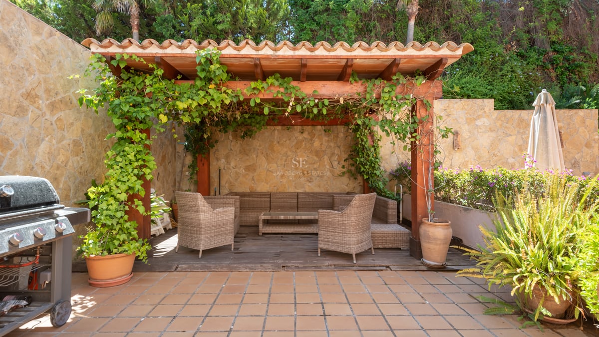 Terrasse extérieure avec pergola en bois, meubles en osier, murs en pierre et barbecue entouré de verdure.