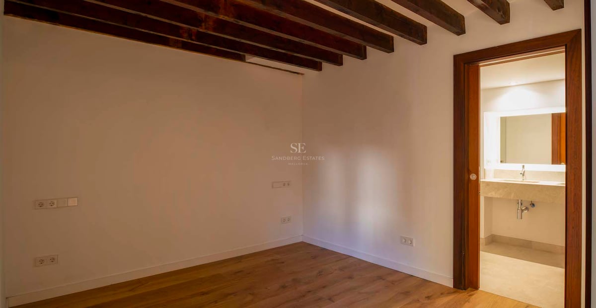 Chambre vide avec poutres de plafond en bois foncé, parquet et porte ouvrant sur une salle de bain moderne.