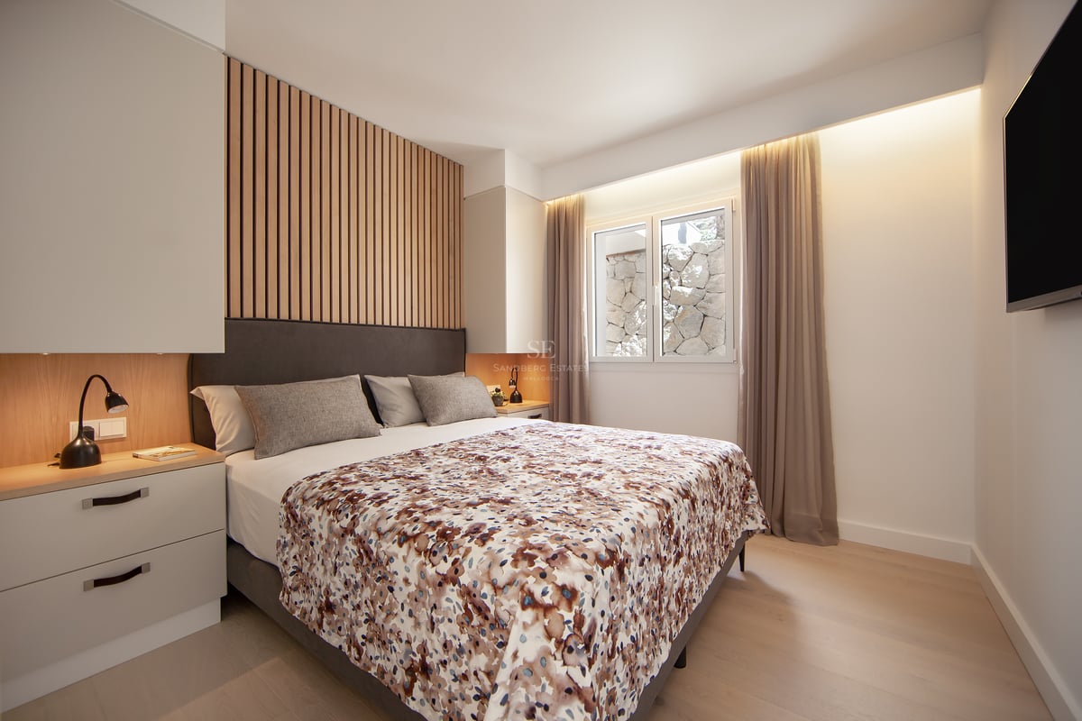 Chambre moderne avec lit gris, détail de mur en bois, éclairage chaleureux et fenêtre avec rideaux.