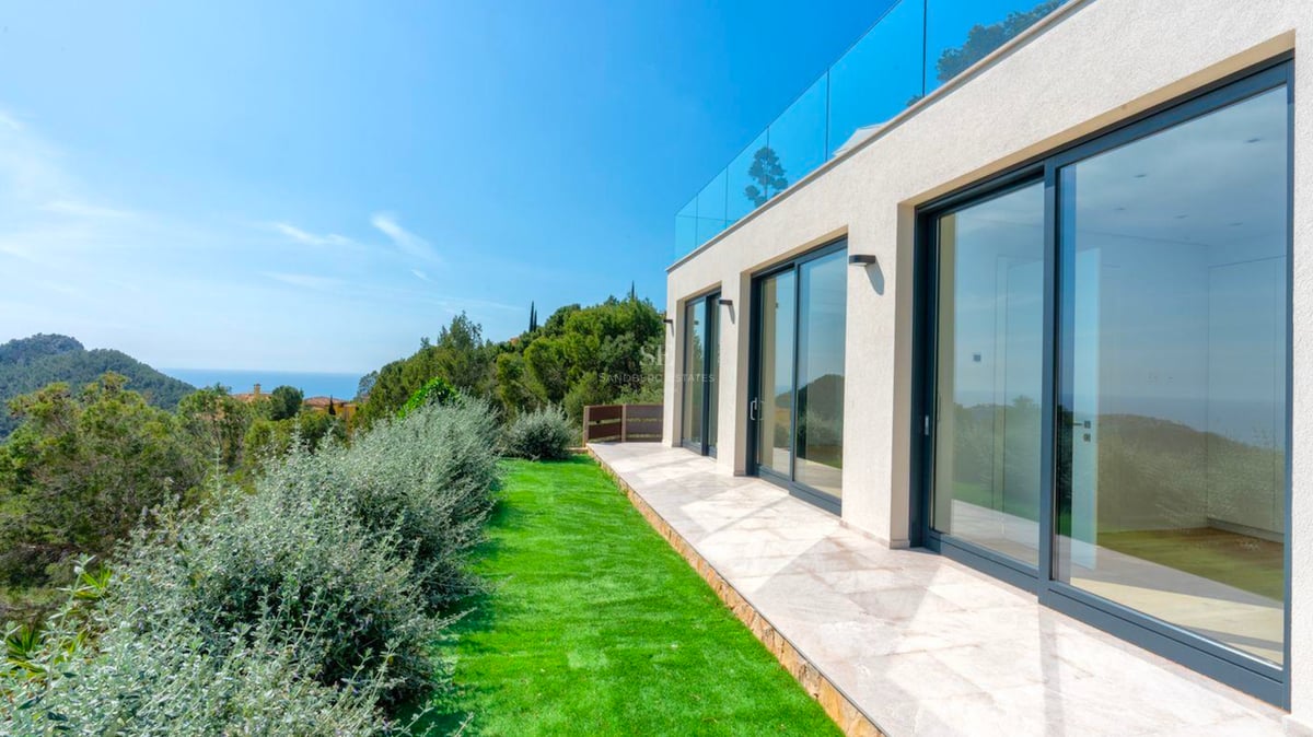 Villa blanche moderne avec grandes portes vitrées coulissantes, patio en pierre, pelouse et vue sur la mer et les montagnes.