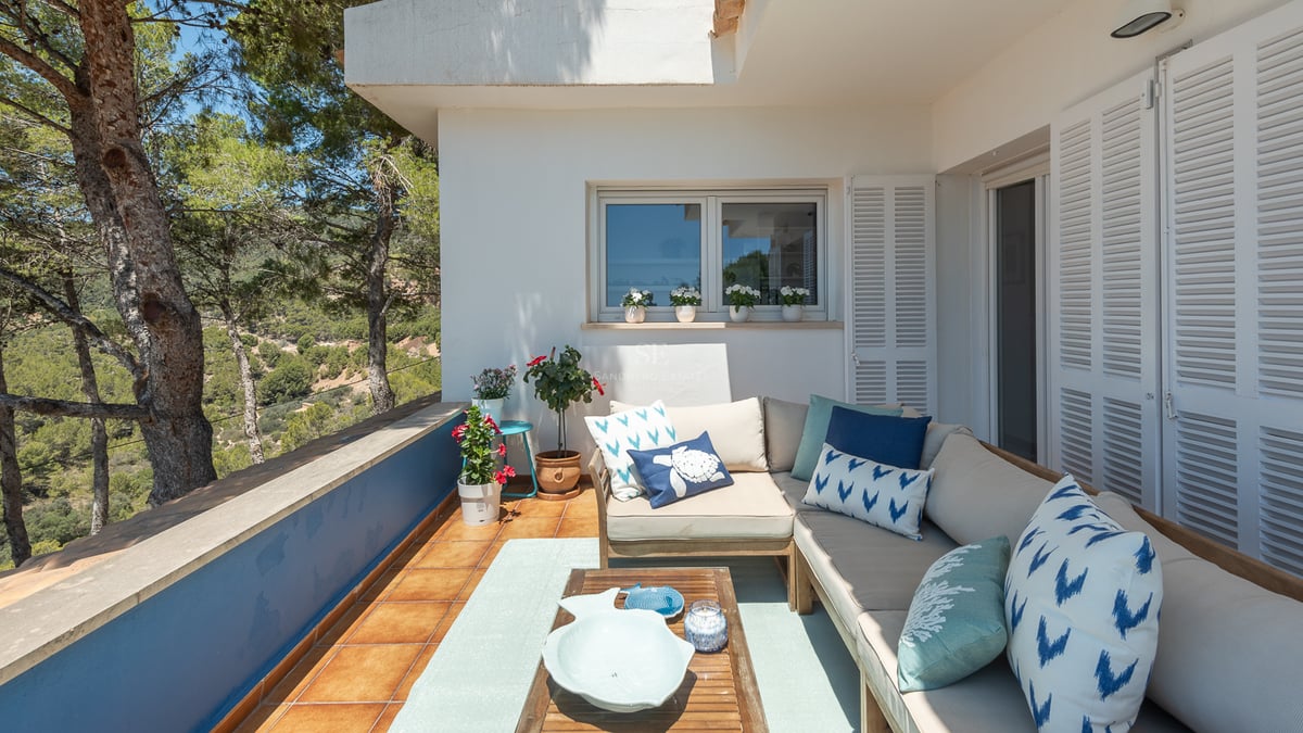 Terrasse extérieure avec un grand canapé en L, des coussins bleus et une table en bois surplombant une vallée de pins.