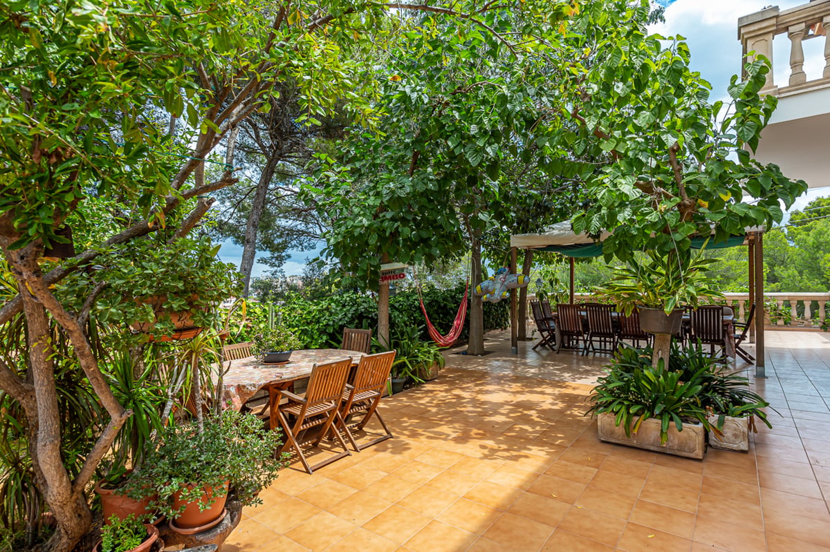 Grande terrasse carrelée entourée d'arbres matures avec deux espaces repas en bois et un hamac rouge à l'ombre.