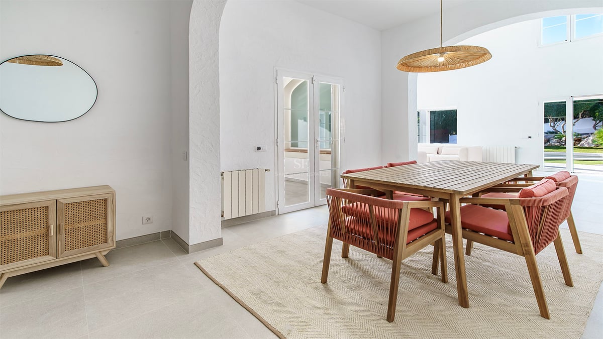 Salle à manger lumineuse avec table en bois, chaises terre cuite, luminaire en osier et arches blanches.
