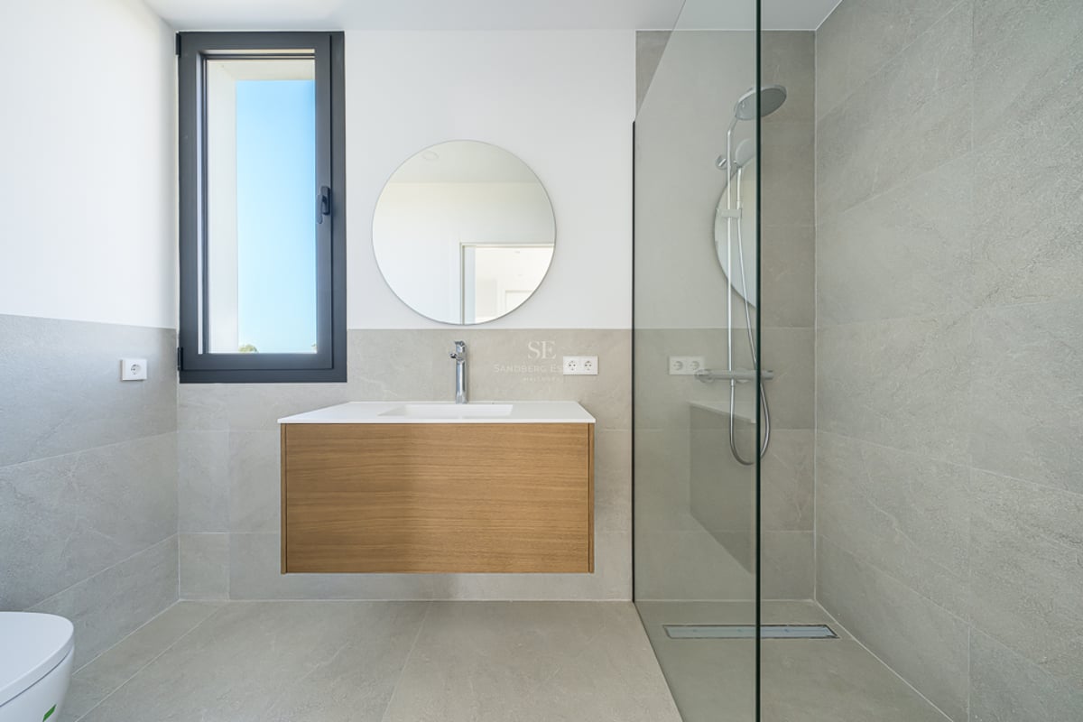 Salle de bain moderne avec carrelage gris, meuble en bois suspendu sous un miroir rond et douche à l'italienne vitrée.