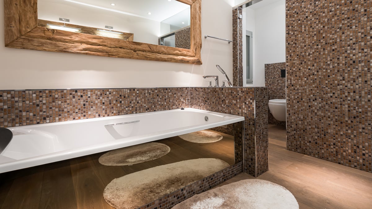 Baignoire blanche moderne avec base en miroir, mur en mosaïque marron et grand miroir avec cadre en bois brut.