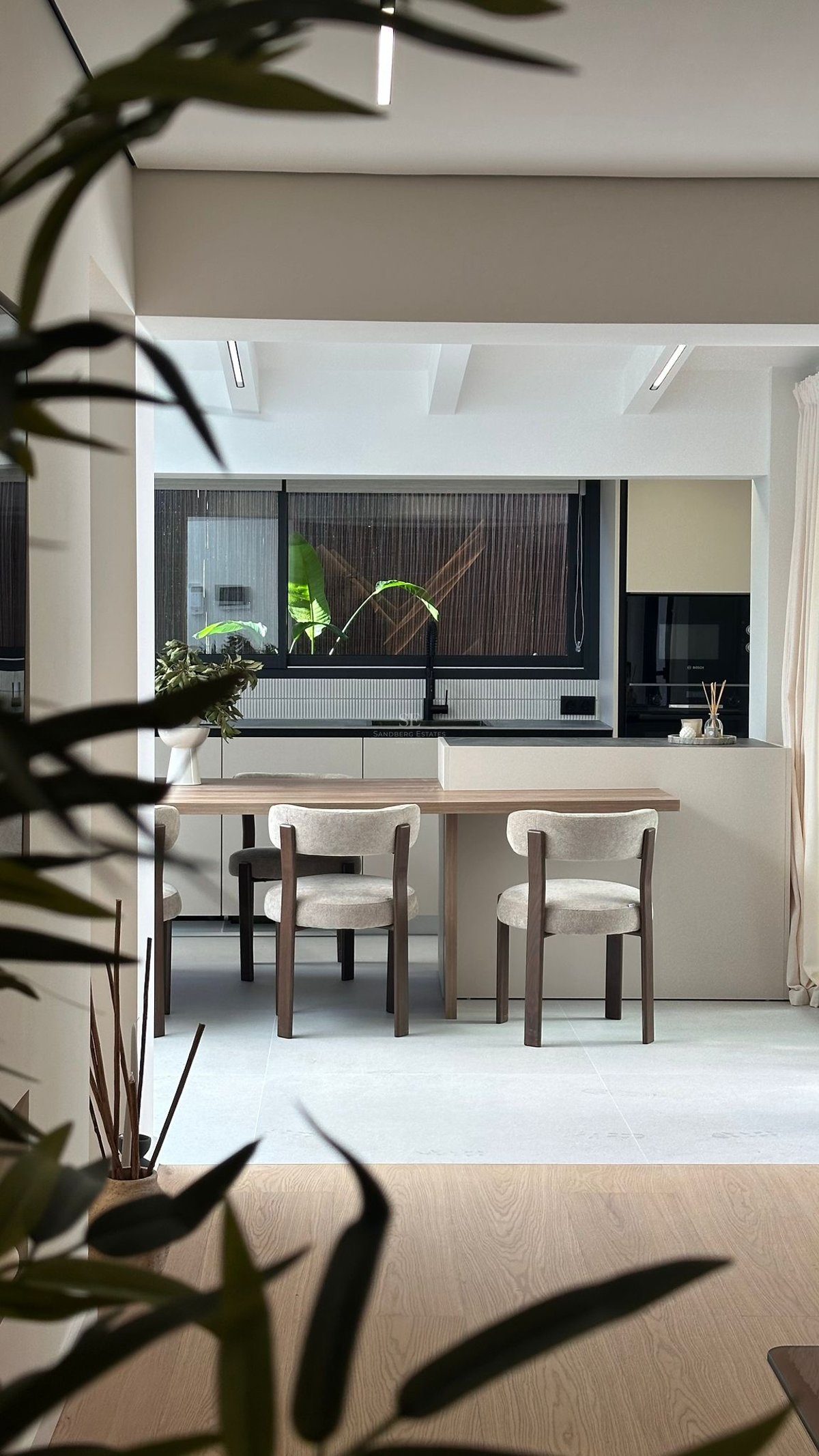 Cuisine moderne avec table en bois intégrée à un îlot beige, chaises design et grande fenêtre.