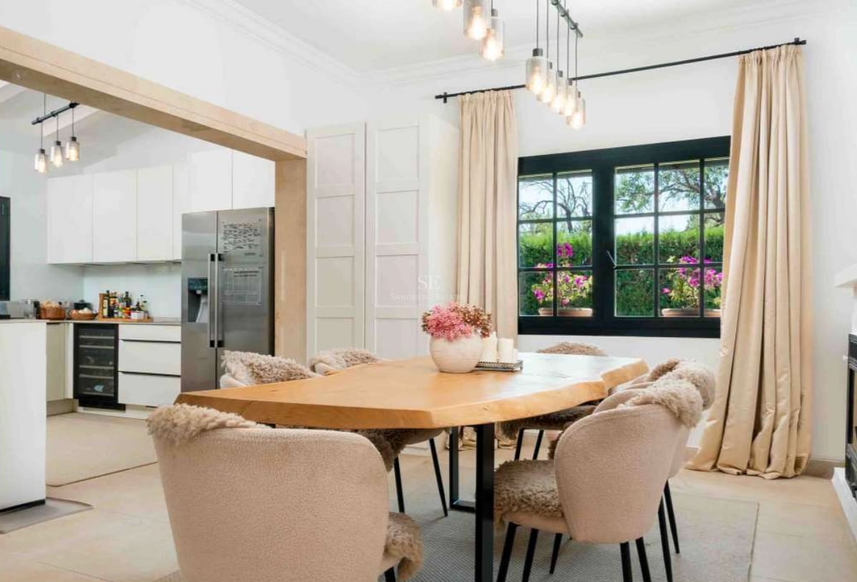 Salle à manger avec table en bois massif, chaises beiges et vue sur une cuisine blanche moderne avec cave à vin.