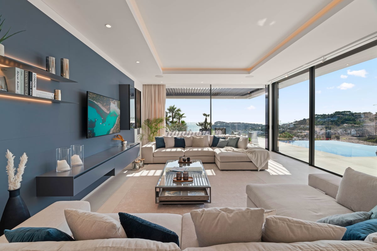 Vaste salon moderne avec canapés beiges, mur d'accent bleu et baies vitrées donnant sur la mer et une piscine.