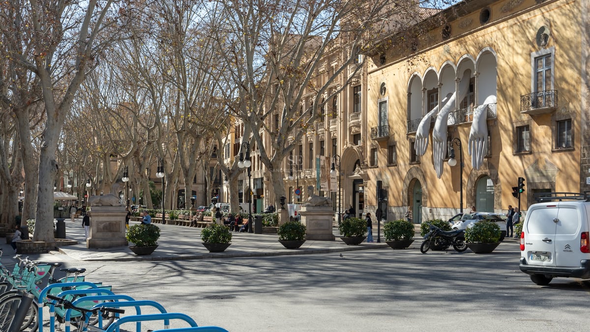 A tree-lined urban promenade with historic Mediterranean buildings, stone statues, and a sculpture of giant white hands.