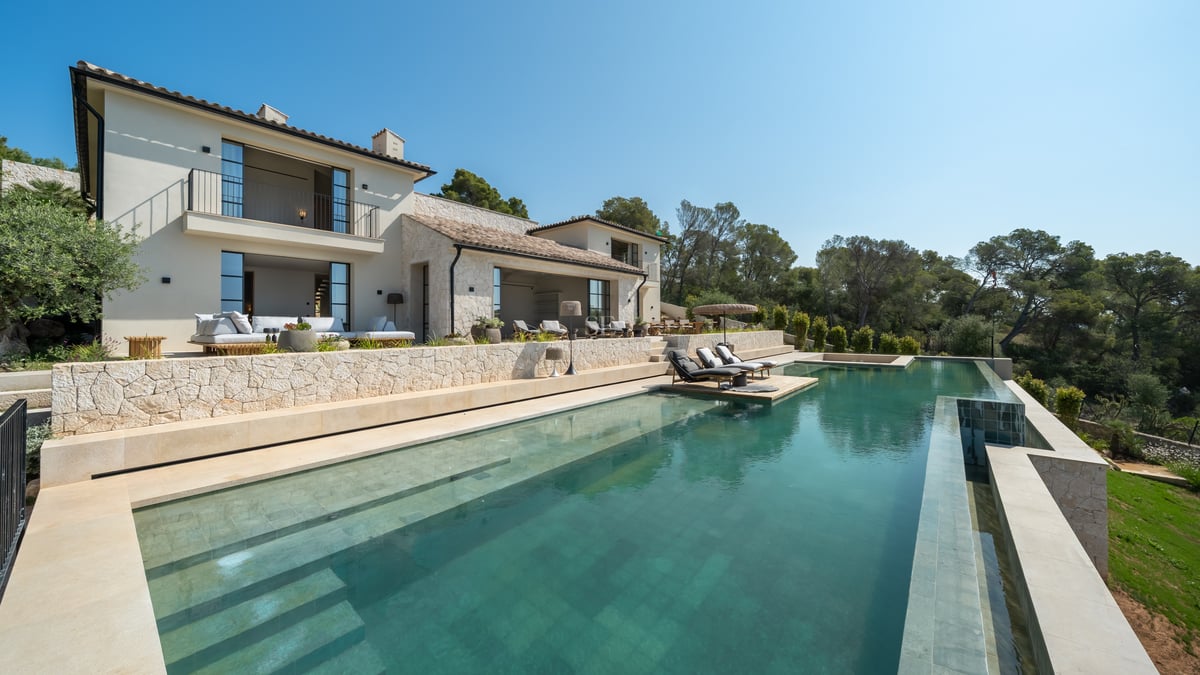 Vue d'une villa de luxe avec une grande piscine à débordement, murs en pierre et bains de soleil face à la forêt.