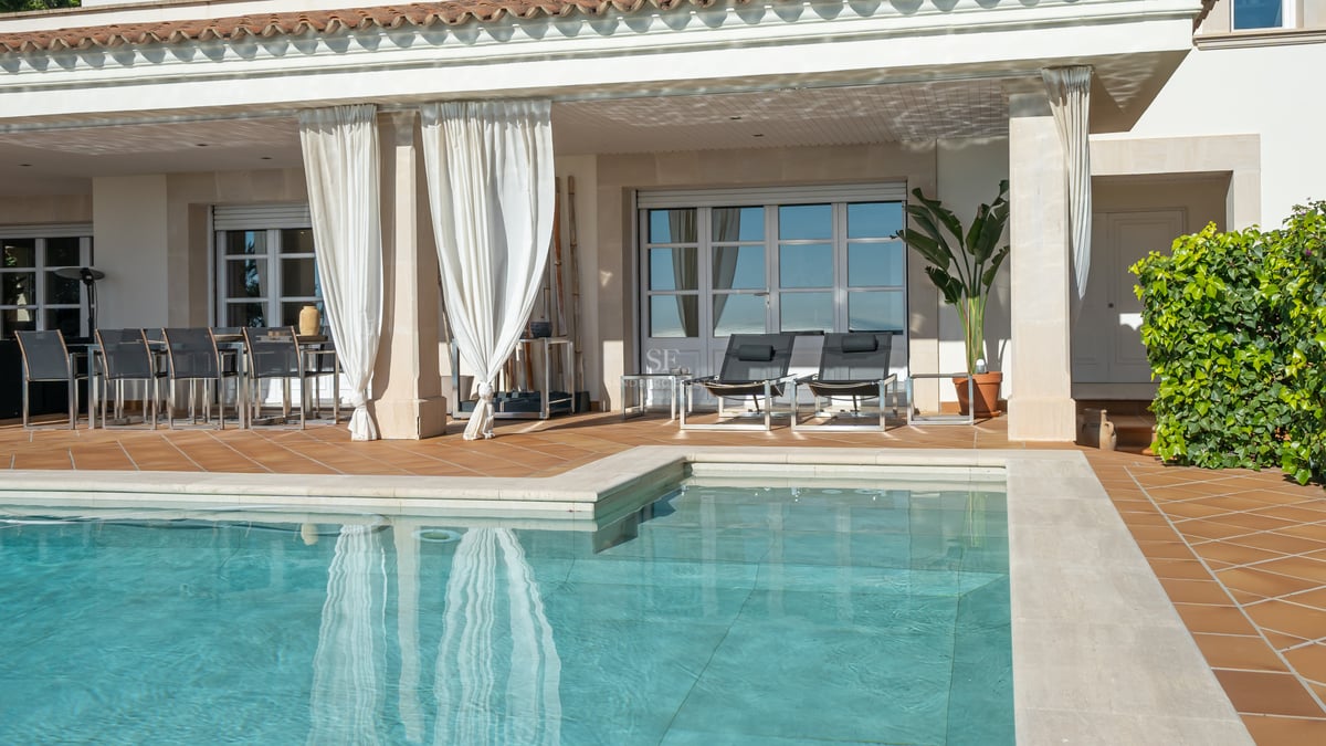 Terrasse en terre cuite avec piscine, rideaux blancs, chaises longues et coin repas extérieur abrité.